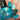 A close-up of a hand with teal nail polish applied, holding a bottle of teal nail polish. The nails are styled with white and black floral patterns, and the bottle label reads 'Hit the Bottle!'.