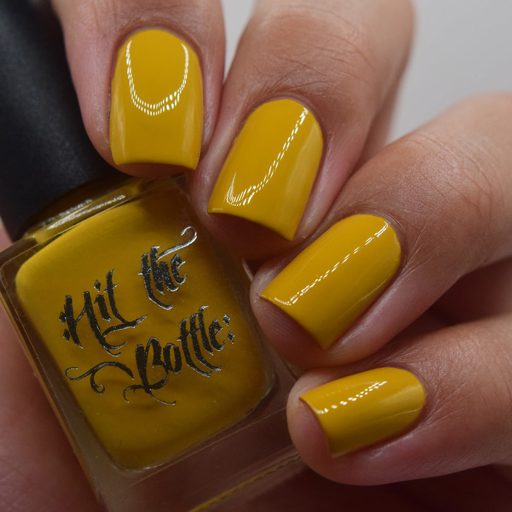 Hand with mustard yellow nails holding a bottle of 'Hit the Bottle' nail polish.